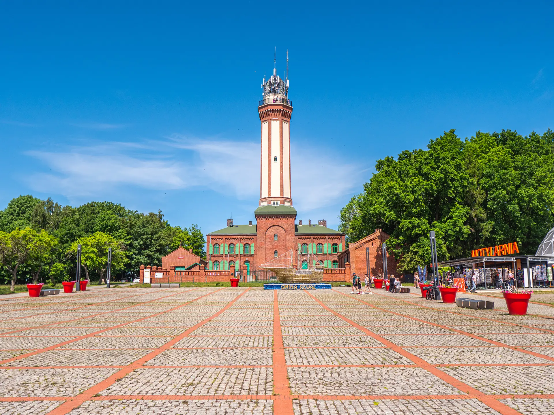 Latarnia morska w Niechorzu nad ceglaną zabudową i brukowanym dziedzińcem z donicami